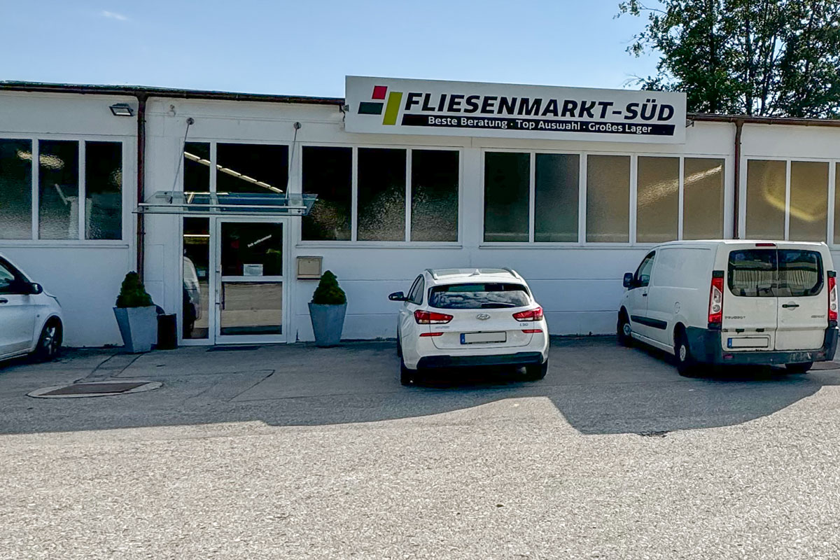 Außenansicht des Fliesenmarkts Süd in Geretsried. Der Eingang des Gebäudes ist mit einem modernen Glasvordach ausgestattet. Über dem Eingang prangt ein großes Schild mit dem Logo des Fliesenmarkts und dem Slogan „Beste Beratung · Top Auswahl · Großes Lager“. Vor dem Gebäude befinden sich zwei parkende Fahrzeuge und zwei dekorative Pflanzkübel, die den Eingangsbereich einrahmen. Die große Fensterfront bietet Einblick in die Innenräume und vermittelt einen einladenden Eindruck. Ein sonniger Tag unterstreicht die freundliche Atmosphäre des Standorts.