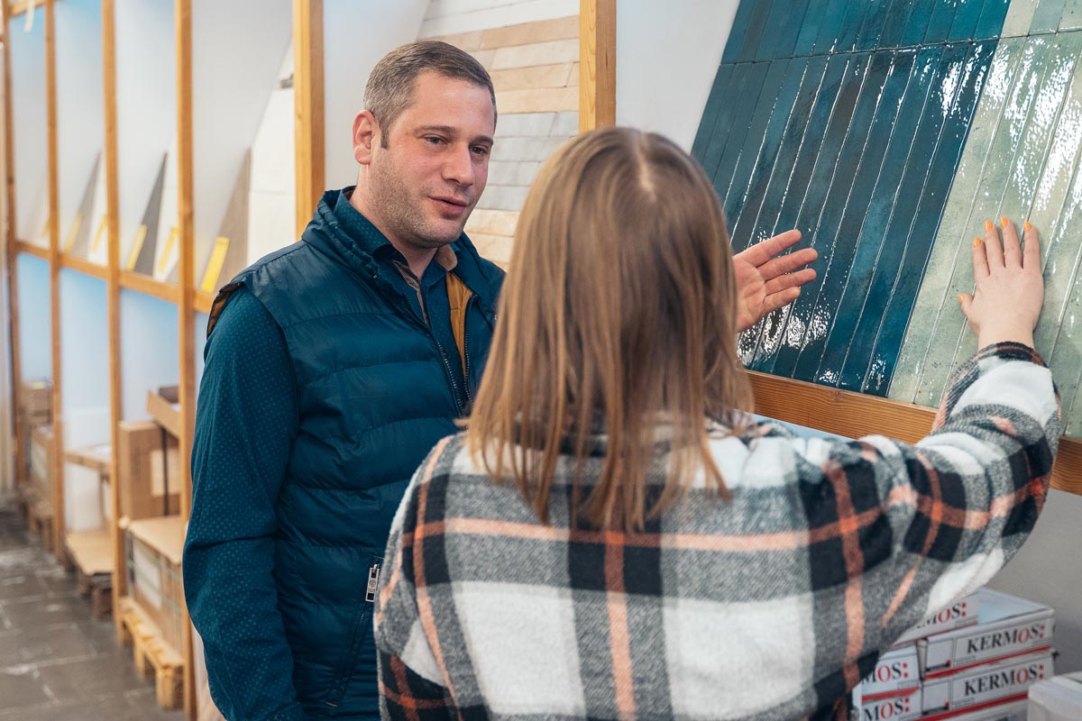 Matthias Lanzl, Geschäftsführer des Fliesenmarkt-Süd, berät eine Kundin vor einer Auswahl hochwertiger Fliesen. Im Fokus stehen glänzende, blaue Fliesenmuster, die stilvoll präsentiert werden. Die Szene unterstreicht die persönliche Beratungskompetenz und die vielfältige Produktpalette des Fachmarkts.