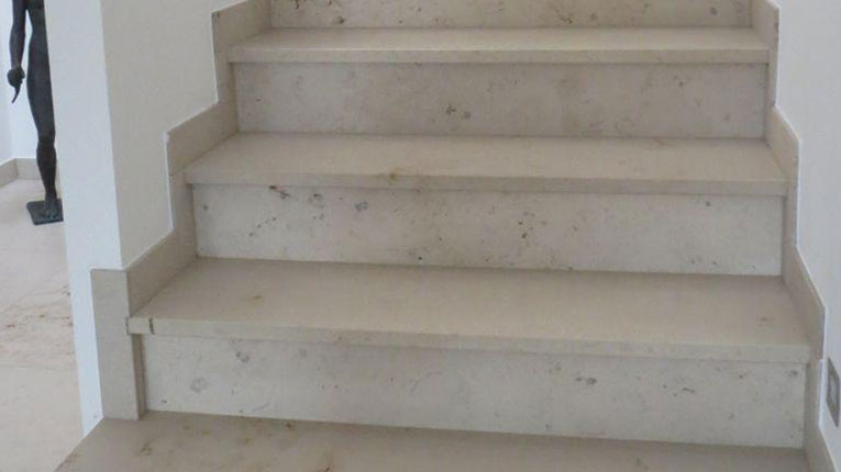 Eine innenliegende Treppe mit Stufen aus hellem Naturstein. Die glatten Oberflächen und die präzise Verarbeitung der Stufen schaffen ein hochwertiges und modernes Erscheinungsbild. Die Seiten der Stufen sind passend verkleidet, um einen einheitlichen Look zu gewährleisten. Der Naturstein zeigt dezente Maserungen und sorgt für eine natürliche Ästhetik. Im Hintergrund ist ein Teil des Flurbereichs und eine Skulptur zu erkennen, was den Raum stilvoll ergänzt. Die Treppe eignet sich ideal für eine langlebige und elegante Gestaltung von Innenräumen.