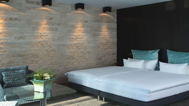 Elegantes Hotelzimmer mit einer Kombination aus modernem Design und natürlichen Elementen. Die Rückwand des Bettes ist mit stilvoll beleuchtetem Naturstein verkleidet, wodurch ein gemütliches Ambiente entsteht. Das Doppelbett ist mit frischen weißen Bettbezügen und türkisfarbenen Kissen ausgestattet. Im Vordergrund befindet sich eine Sitzgelegenheit mit einem modernen Glastisch, auf dem eine dekorative Blumenschale steht. Die dunklen Akzente der Möbel und der Teppichboden kontrastieren harmonisch mit den hellen Wandfarben.