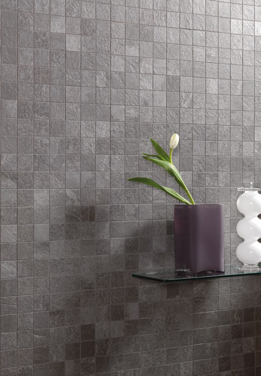 Eine Wand mit kleinen quadratischen Mosaikfliesen in dunklem Anthrazitgrau, die durch ihre dezente Struktur und matte Oberfläche einen eleganten, zeitlosen Look erzeugen. Auf einem schwebenden Glastablar stehen eine minimalistische, purpurfarbene Vase mit einer einzelnen weißen Blume und eine moderne, weiße Design-Dekoration. Perfekt für ein modernes Wohn- oder Badezimmerambiente mit luxuriösem Touch.