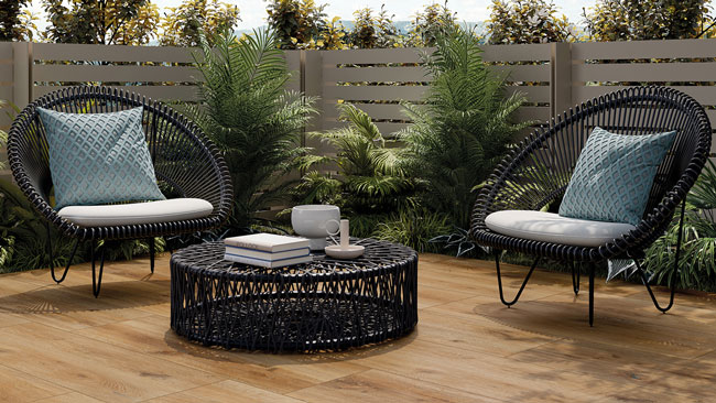 Modern gestaltete Terrasse mit Fliesen in realistischer Holzoptik, die eine warme und einladende Atmosphäre schaffen. Zwei schwarze Rattan-Sessel mit hellen Sitzpolstern und gemusterten blauen Kissen rahmen einen runden Rattan-Couchtisch ein. Der Hintergrund zeigt eine Sichtschutzwand in Beige und eine üppige Begrünung mit Farnen und anderen Pflanzen. Die Fliesen bieten eine langlebige und pflegeleichte Alternative zu echtem Holz und sind ideal für den Außenbereich.